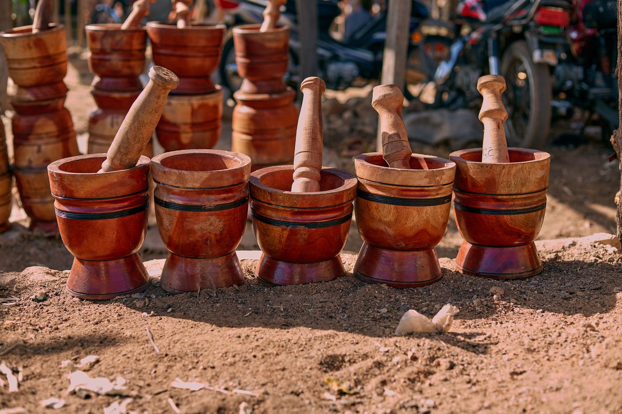 about-img A lineup of handcrafted wooden mortars and pestles displayed on a sunny outdoor market day.